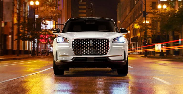 White SUV with illuminated grille driving through a city street at night, with buildings and lights in the background.