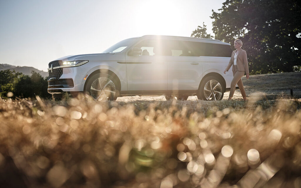 A person walking beside a white SUV in a sunlit field, with a tree and hills in the background.