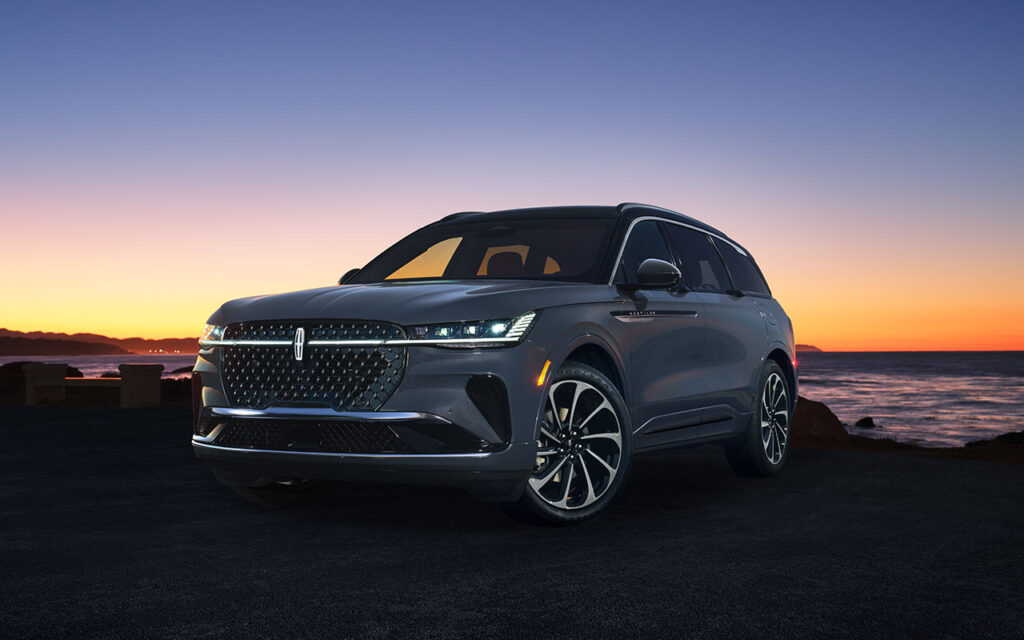Luxury SUV parked at sunset by the ocean, featuring sleek design and illuminated headlights.