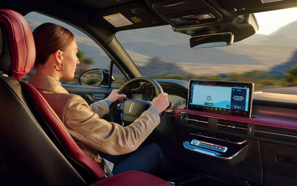 Woman driving vehicle with navigation system on screen, surrounded by scenic landscape.