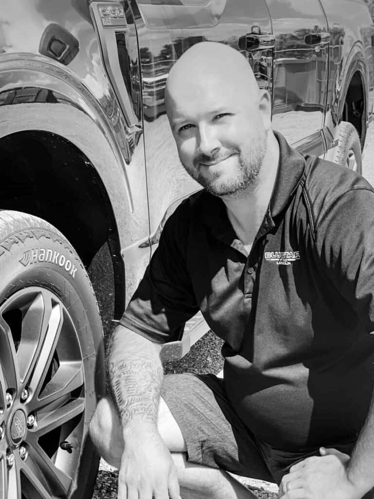 Smiling man kneeling by a car, wearing a black polo shirt with "Bluestar" logo, black and white.